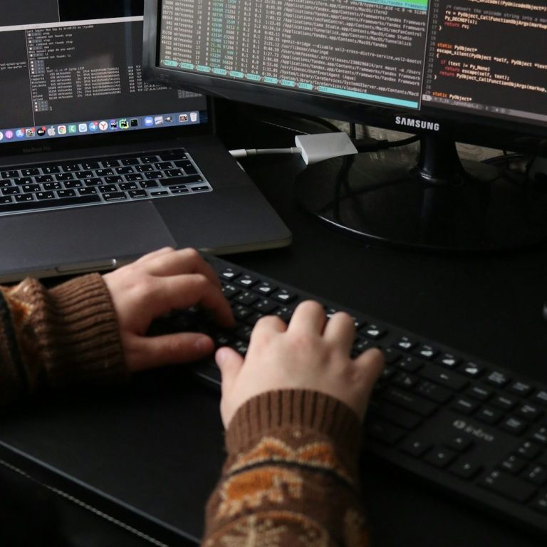 Hände tippen auf einer Tastatur vor mehreren Monitoren mit Code und Daten.